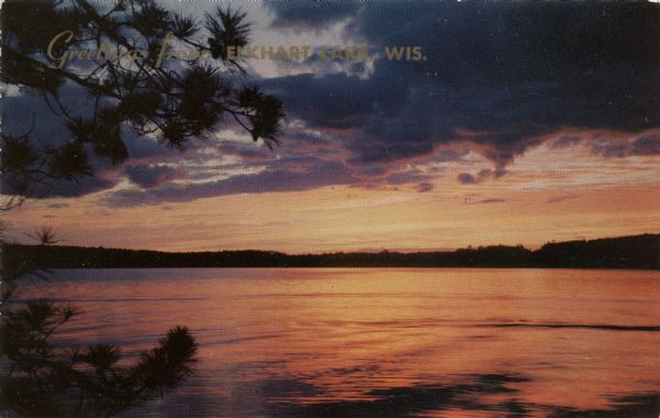 Kodachrome print of twilight over Elkhart Lake, with purple clouds and an orange sky. Caption reads: "Greetings from Elkhart Lake, Wis." Text on reverse reads: "Shimmering waters The sun sets gloriously at the end of another perfect day. Vacationland scene."