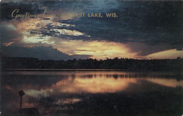 Sunset over Elkhart Lake, with the calm lake reflecting the orange sky. Caption reads: "Greetings from Elkhart Lake, Wis." Text on reverse reads: "Sunset at the end of a perfect day. Vacationland scene."