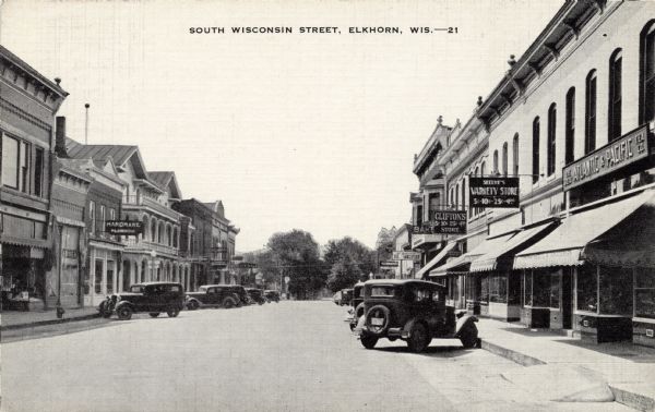 Street scene in a commercial district lined with shops and street lamps. Automobiles are parked at an angle to the curbs on both sides of the street. Caption reads: "South Wisconsin Street, Elkhorn, Wis."