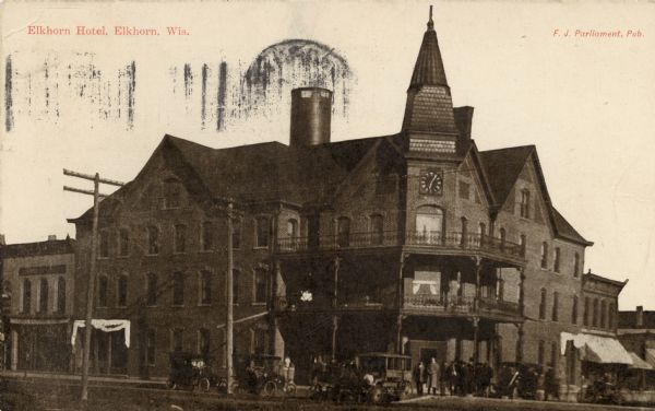 View of a large, brick corner hotel with balconies on the second and third floors, and a clock above the entrance. Men are standing in front. and automobiles are parked in the street. Caption reads: "Elkhorn Hotel, Elkhorn, Wis."