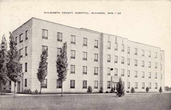 Black and white photographic postcard of the county hospital, a four-story stone building. Young trees and bushes are on the lawn. Caption reads: "Walworth County Hospital, Elkhorn, Wis."