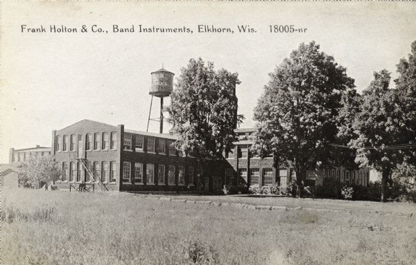 View across field towards a factory where band instruments are manufactured. A water tower with "Holton Band Instruments" painted on it is in the background. Caption reads: "Frank Holton & Co., Band Instruments, Elkhorn, Wis."