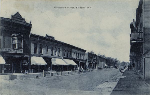 Angled view down sidewalk on right toward the left side of Wisconsin Street which is lined with businesses and hitching posts. Horse-drawn vehicles are moving along the street. Caption reads: "Wisconsin Street, Elkhorn, Wis."