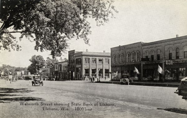 View across Walworth Street. The State Bank of Elkhorn is on the corner with the lamppost. Automobiles are parked in the street. Caption reads: "Walworth Street showing State Bank of Elkhorn, Elkhorn, Wis."