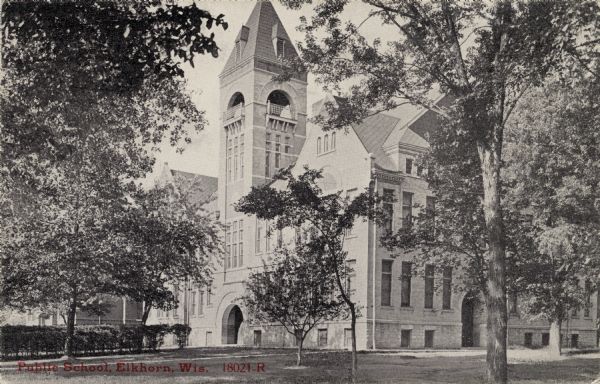 View across lawn toward the public school, which is a large building with a bell tower, arched entrances and gables. Caption reads: "Public School, Elkhorn, Wis."
