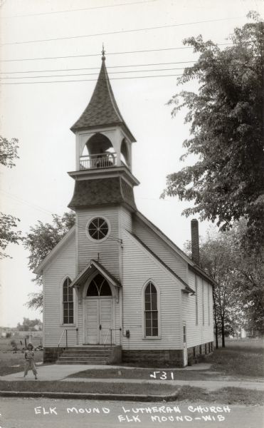 Black and white photographic postcard of a small white church with a bell tower. There is a child standing on the sidewalk in front. Caption reads: "Elk Mound Lutheran Church, Elk Mound — Wis."