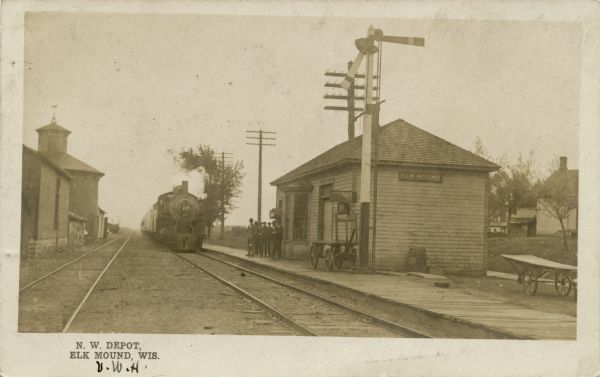 Black and white postcard view of the depot at Elk Mound, with a train approaching. Men are standing on the platform.