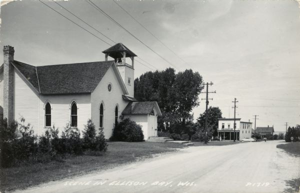 Black and white photographic postcard of a street scene, with a church on the left, and automobiles parked near a storefront at the corner in the distance, with a sign above the entrance that reads: "Volunteer". Caption reads: "Scene in Ellison Bay, Wis."