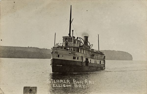 View of the Steamer "Bon Ami" on Ellison Bay. A few people are on the deck. The tree-lined far shoreline is in the background. Caption reads: "Steamer Bon Ami, Ellison Bay, Wis."