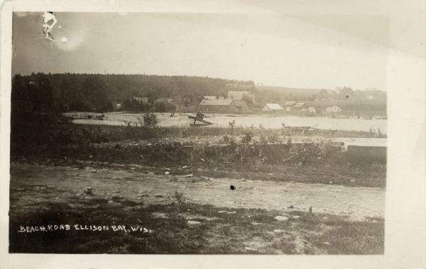 View of Beach Road and the beach at Ellison Bay. Buildings are across the bay, and a tree-lined ridge is in the far background. Caption reads: "Beach Road, Ellison Bay, Wis."