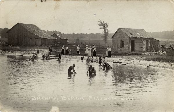 View towards shoreline with a group of people gathered at a bathing beach. Some people are in the water; other people are standing on the shore fully dressed. Caption reads: "Bathing Beach, Ellison Bay."