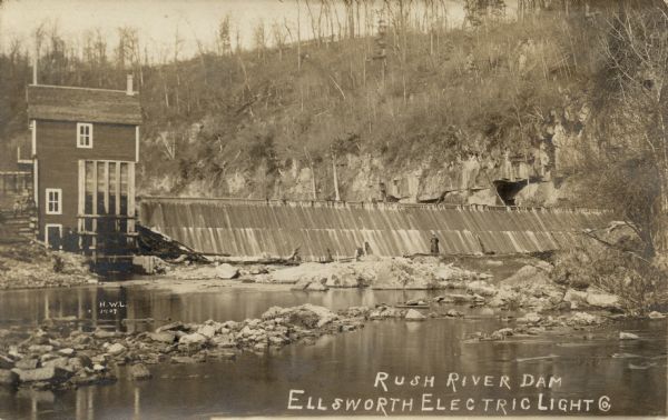 View upriver towards the Rush River Dam. A man is standing on a rock in the middle of the river just below the dam; another man is sitting next to the power house. A high cliff is above the dam in the background. Caption reads: "Rush River Dam, Ellsworth Electric Light Co."