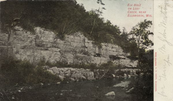 View across stream towards an exposed outcropping at the base of a hill. Caption reads: "Elk Hole on Lost Creek, near Ellsworth, Wis."