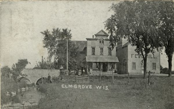 View along stream towards people standing in front of buildings and near a stone bridge. The buildings include a post office and a feed mill. Caption reads: "Elm-Grove, Wis."