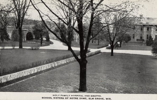 Slightly elevated black and white view of a stone hospital and grotto affiliated with the School Sisters of Notre Dame. Trees and shrubs are on the lawns divided by sidewalks. Caption reads: "Holy Family Hospital and Grotto, School Sisters of Notre Dame, Elm Grove, Wis."