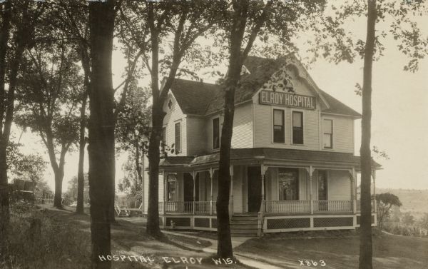 Exterior view of the Elroy Hospital. The two-story building has a wrap-around porch. In the background is a field with piles of harvested grain. Caption reads: "Hospital, Elroy, Wis."