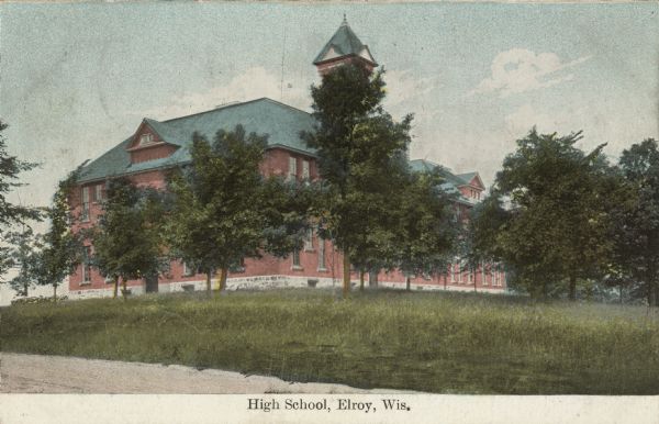 Hand-colored postcard of the high school obscured by trees. The red brick building has two-stories with a bell tower. Caption reads: "High School, Elroy, Wis."
