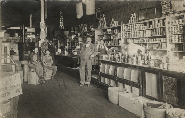 Interior view of a general store with canned goods on display on shelves along the wall in the background. Two men are standing behind the counter, and four men are sitting or standing on the left. Flags are hanging from the ceiling.