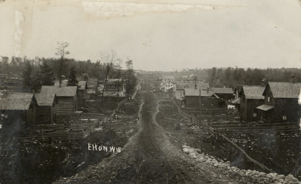 View looking down center of unpaved main street. The street is lined with dwellings with fenced-in yards. Caption reads: "Elton, Wis."