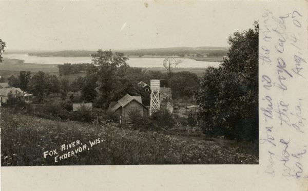 Elevated view from hill towards the Fox River, with a farm in the foreground. The farm has a windmill with a sign on it that reads: "Kilbourn Steel Mill," which is next to a water tower. Caption reads: "Fox River, Endeavor, Wis."