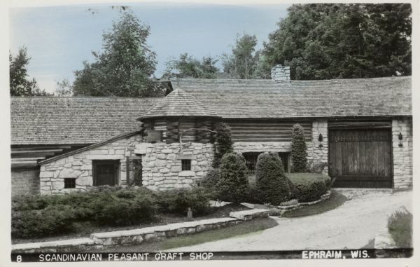 View of the stone building with a large wooden door. There is a driveway lined with a low stone wall with bushes. Caption reads: "Scandinavian Peasant Craft Shop, Ephraim, Wis."