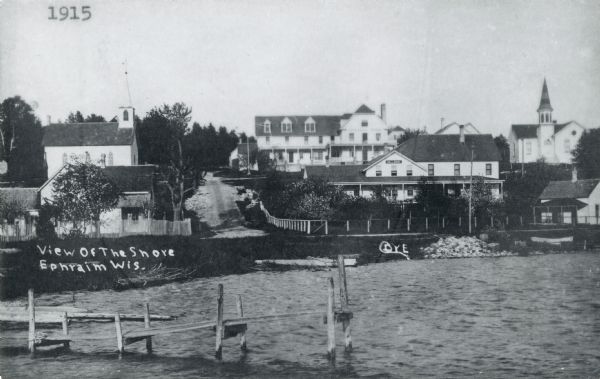 View of Ephraim from the bay, with a pier in the foreground. Churches are on the left and right, and the Hillside Hotel is in the center near the shore. Caption reads: "View of the Shore, Ephraim, Wis."