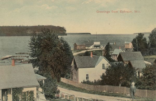 Elevated view from hill of the bay and Ephraim. A man is walking up the hill on a road with buildings and fences on both sides. Caption reads: "Greetings from Ephraim, Wis."