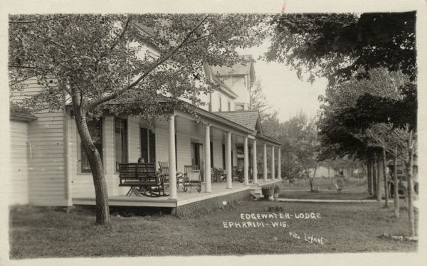 View across lawn towards the Edgewater Lodge, a hotel with a columned porch with a swing and rocking chairs. Caption reads: "Edgewater-Lodge, Ephraim-Wis."