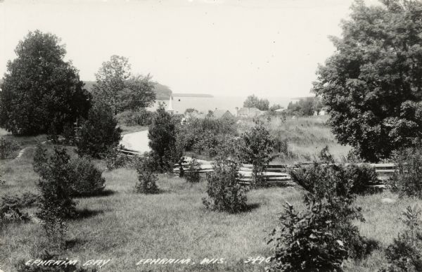 Photographic postcard view of Ephraim and bay from a grassy lot with trees, shrubs and a fence. Caption reads: "Ephraim Bay, Ephraim, Wis."