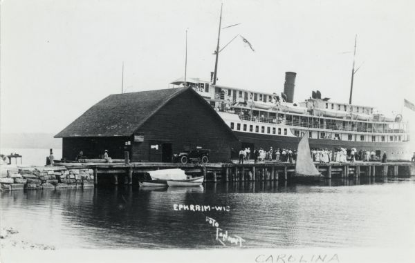 View across water towards the "Carolina" steamer docked at Ephraim. Many people are standing on the pier. Caption reads: "Ephraim-Wis."