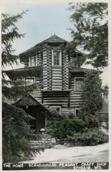 Hand-colored black and white photographic postcard of a three-story log structure, and roofed entrance. Caption reads: "The Home; Scandinavian Peasant Craft Shop, Ephraim, Wis."