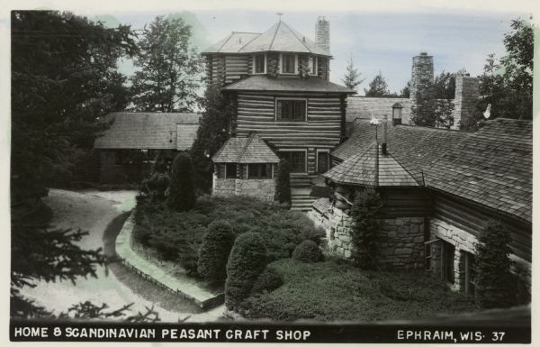 Hand-colored black and white photographic postcard with an elevated view of the three-story log structure and attached buildings with shrubs in the yard, and a low stone wall along the drive. Caption reads: "Home 8, Scandinavian Peasant Craft Shop, Ephraim, Wis."