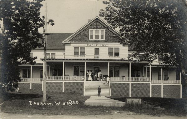 The Eagle Inn, a two-story wooden structure with a gabled roof, balcony and porch. Women and children are gathered on the front steps. A dog is running in the yard on the left. Caption reads: "Ephraim, Wis."