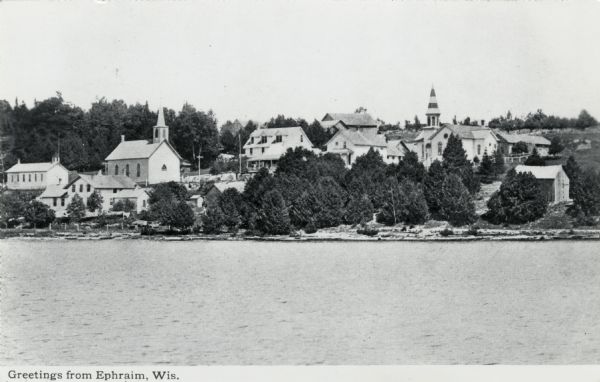 Black and white photographic postcard view of Ephraim from Eagle Bay. Churches and dwellings are on the hill above the shoreline. Caption reads: "Greetings from Ephraim, Wis."