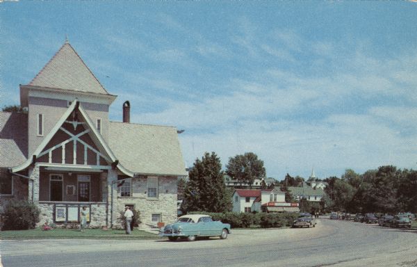 View across street towards the Ephraim Village Hall. Automobiles are parked on both sides of the street.

Text on reverse reads: "A view of Ephraim's picturesque village hall with its quaint Norwegian architecture showing Wilson's Restaurant and Gift Shop in the background, located 27 miles north of Sturgeon Bay, on Highway 42."