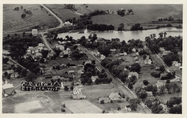 Photographic postcard aerial view of Ettrick. A river is running through town. Dwellings and commercial buildings are surrounded by fields.