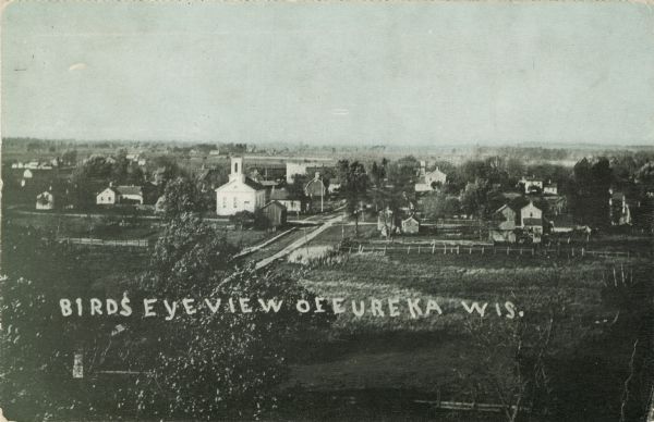 Elevated view of the town, with a church and dwellings, surrounded by fields. Caption reads: "Birds [sic] Eye View of Eureka, Wis."