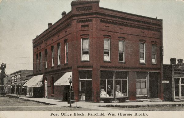 Hand-colored corner view from intersection towards the post office. A postcard display is inside the window. Caption reads: "Post Office Block, Fairchild, Wis. (Burnie Block)." 