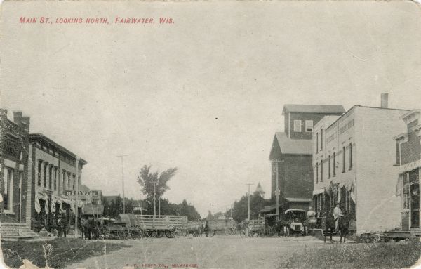 View of the central business district of Fairwater. Horses and wagons and automobiles in the street. A hardware store is on the left. Two men are on the right, looking towards the camera. Caption reads: "Main St., Looking North, Fairwater, Wis."