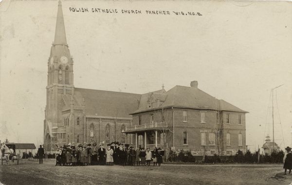 View towards the Polish Catholic Church and parsonage. There is a crowd of women and children standing in the unpaved street in front. Caption reads: "Polish Catholic Church, Fancher, Wis."