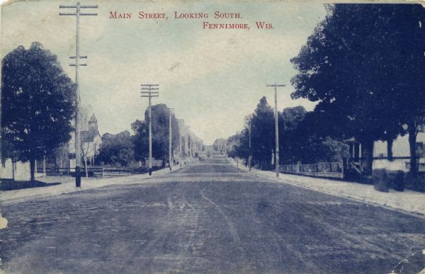 View down center of unpaved Main Street, which is lined with trees in the foreground; the central business district is further down the street. Caption reads: "Main Street, Looking South. Fennimore. Wis."