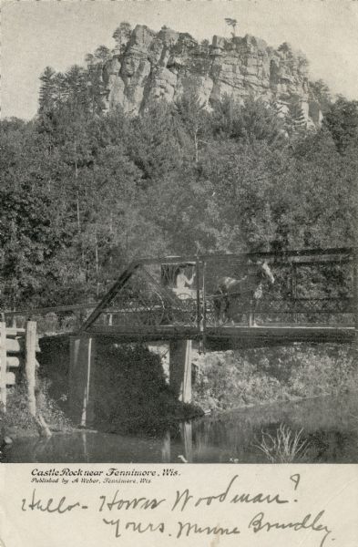 View of Castle Rock in the distance. In the foreground a woman is driving a horse-drawn carriage across a bridge. Caption reads: "Castle Rock near Fennimore, Wis."