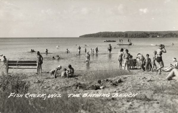 View from shoreline looking out towards the public beach with several bathers. Diving platforms are out in the bay. Caption reads: "Fish Creek, Wis. The Bathing Beach."