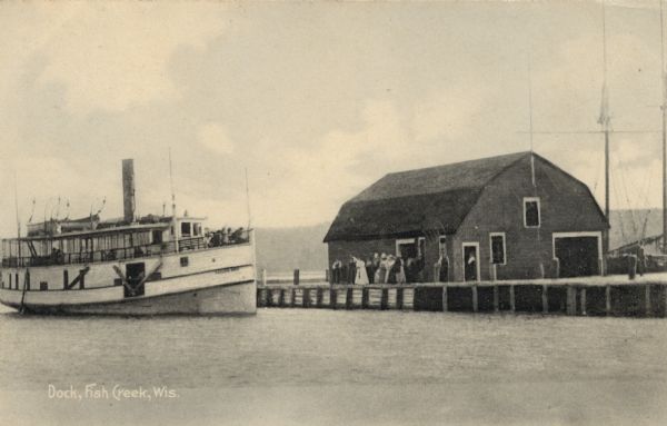 View across water towards the Fish Creek Dock with a boathouse, and a steamer pulling up from the left. A group of people are gathered on the dock. Caption reads: "Dock, Fish Creek, Wis."