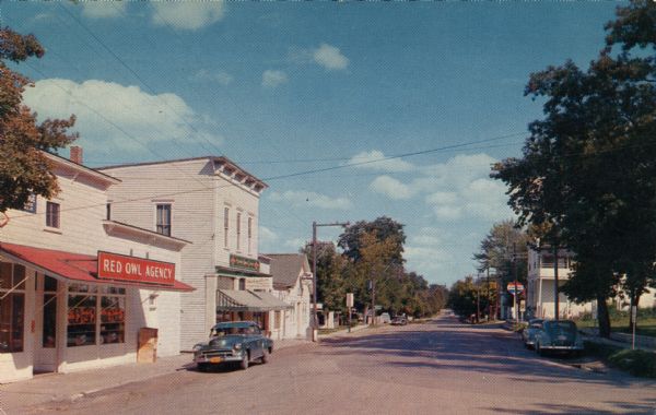 Color print of a view of Main Street. The Red Owl Agency is on the left, and a Standard Service Station is further down the street on the right. Cars are parked along the curb on both sides of the street.