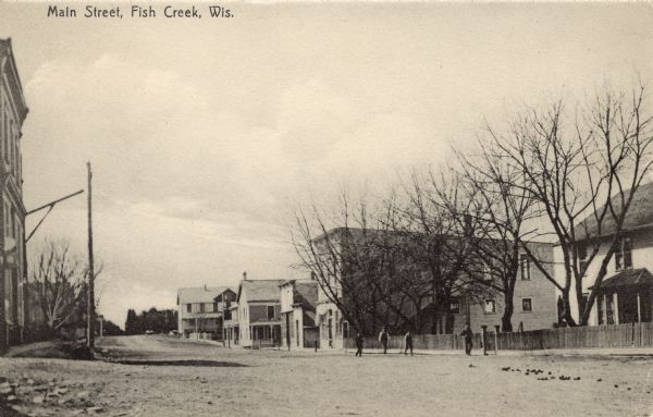 Sepia-toned view down unpaved Main Street. Buildings, dwellings and trees are lining the street. Four men are standing in the street near a fence in the center. Caption reads: "Main Street, Fish Creek, Wis."