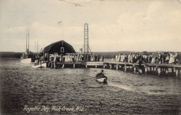 View across water from shoreline towards a crowd of people gathered on the main dock. Boats are in the water near the dock, and one person is rowing a boat near the pier. Caption reads: "Regatta Day, Fish Creek, Wis."