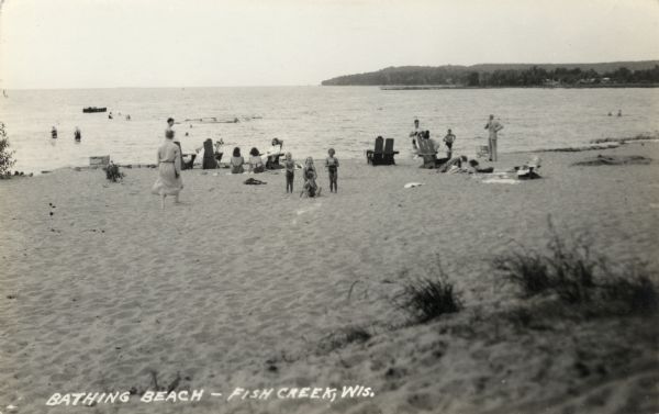 View from dunes towards a beach with bathers. The main dock and Peninsula State Park are in the distance. Caption reads: "Bathing Beach - Fish Creek, Wis."