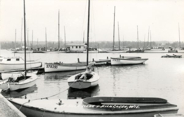 View of the marina, with several docked sailboats. The tree-lined shoreline is in the background. Caption reads: "Busy Pier Scene, Fish Creek, Wis."