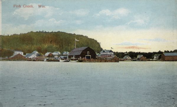Hand-colored view of Fish Creek from the bay. A boathouse and main dock are in the center. A bluff is in the background. Caption reads: "Fish Creek, Wis."